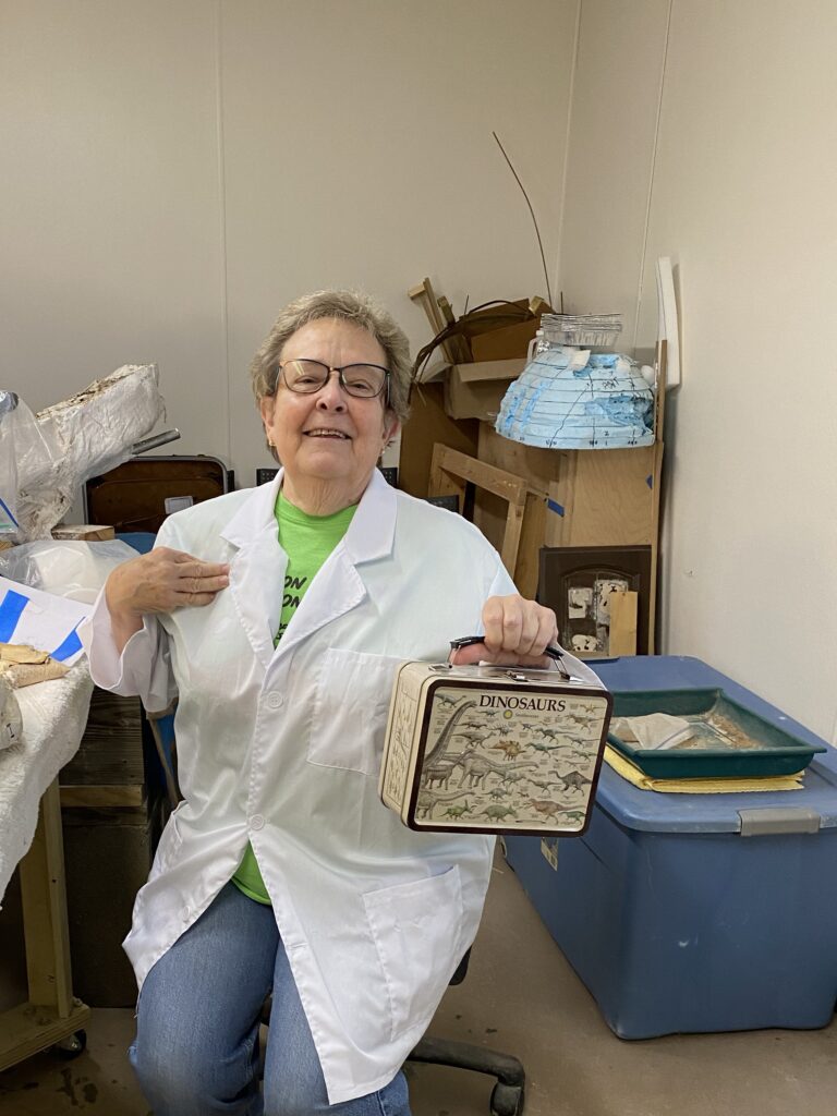 Deborah Canterbury smiling wearing a lab jacket holding her dinosaur lunchbox