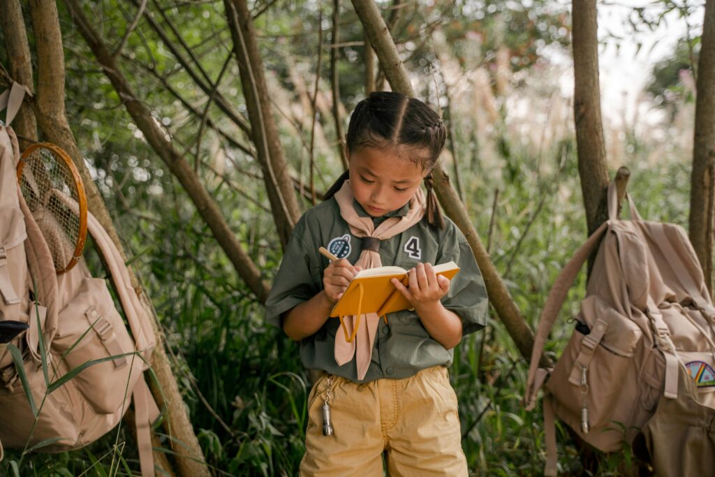 A young girl scout takes notes during a summer camp in the woods.