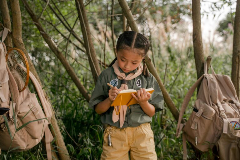 A young girl scout takes notes during a summer camp in the woods.