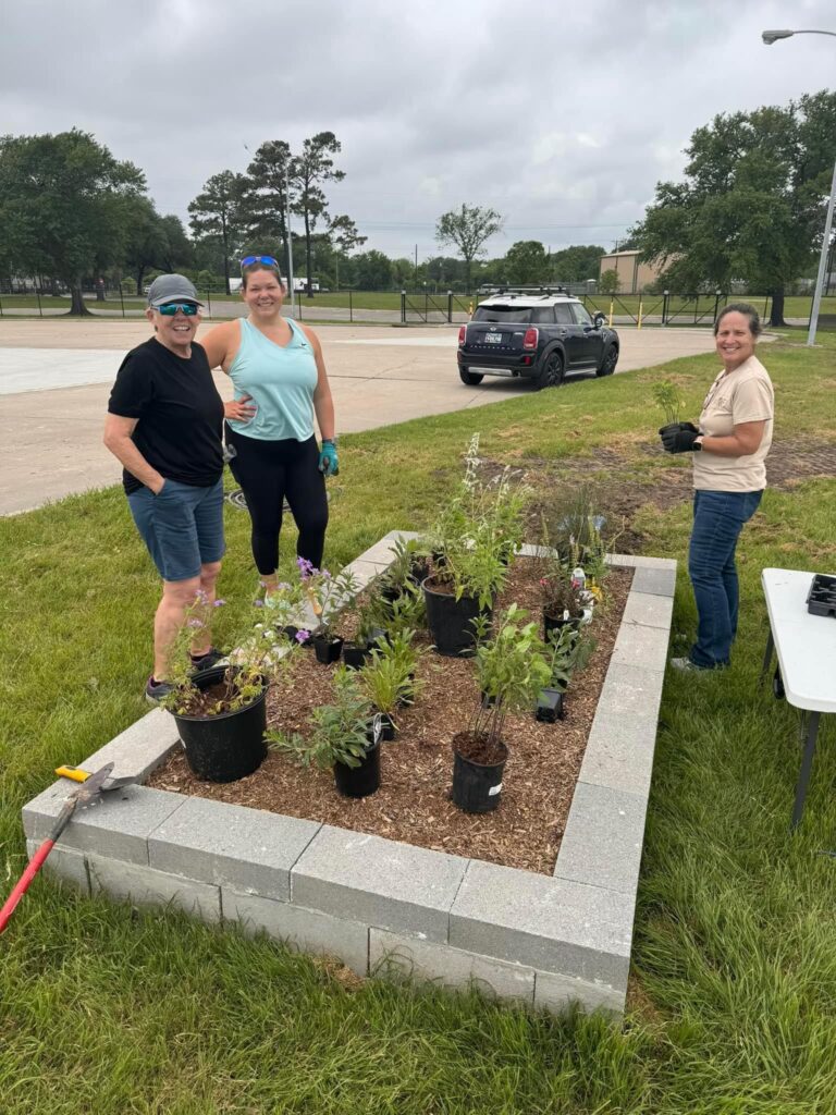 A raised garden bed made of cinder blocks with mulch and various green plants being staged for planting. Three volunteers stand around the garden.